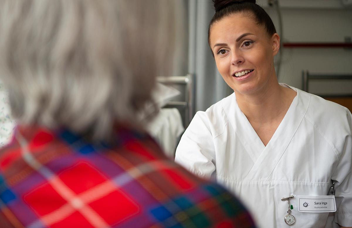 En sykepleier snakker med en person i kofte. Hen med kofte har ryggen til.