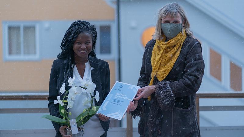 2 damer står utendørs, hvor ene mottar et diplom fra den andre