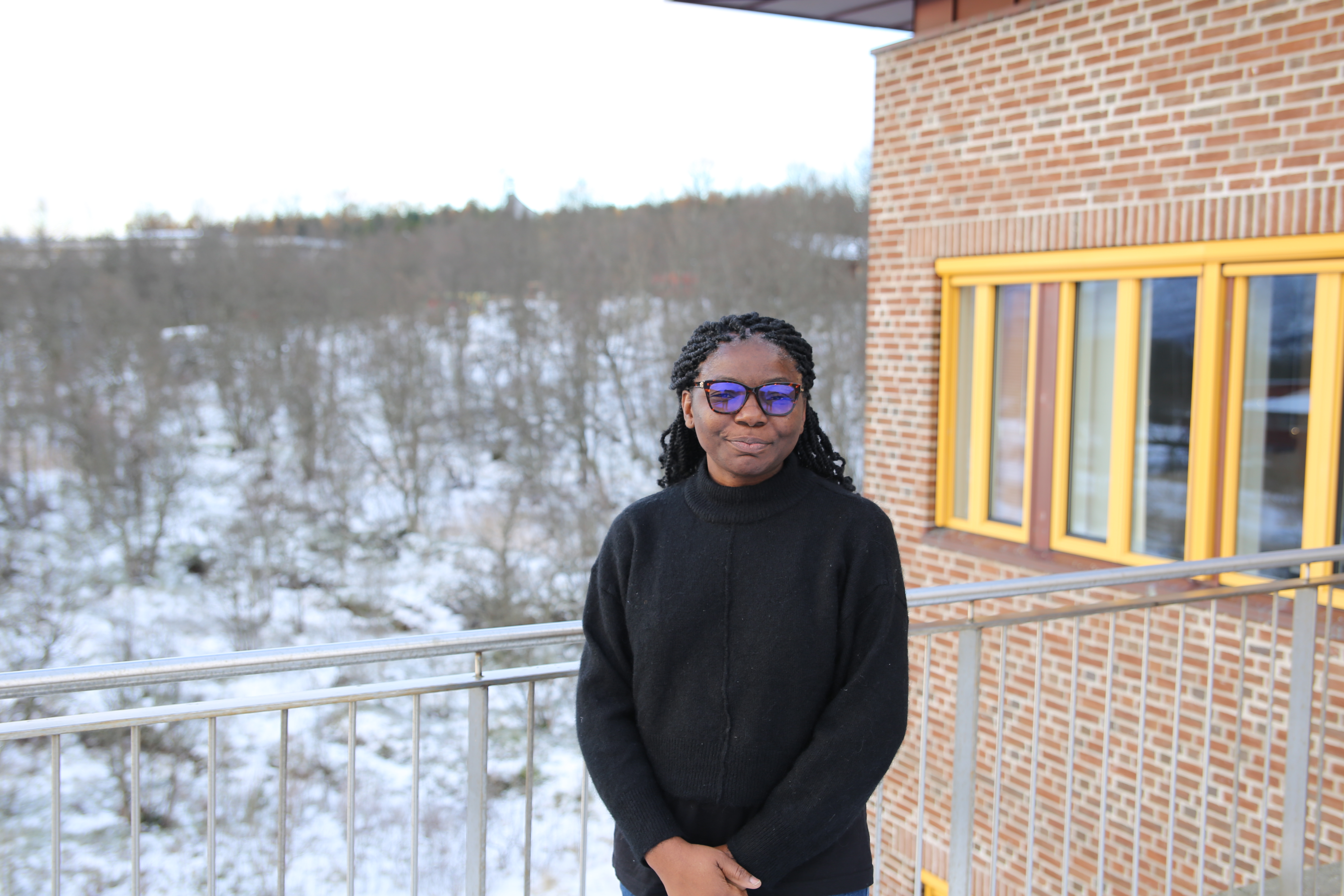 Picture of Sheila standing outside of a building. It is late fall, so there is snow on the trees in the background. 