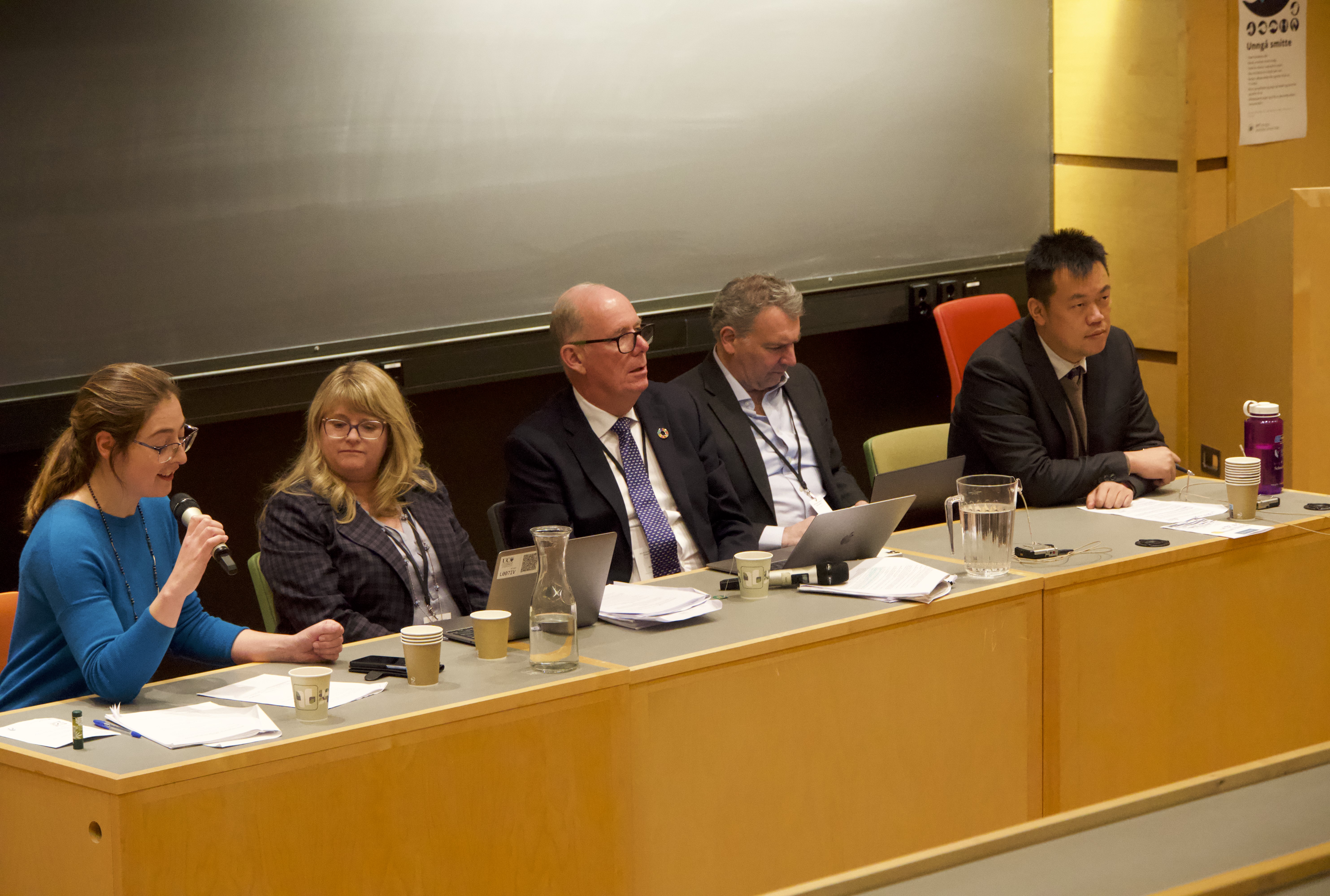 Eva is speaking in a microphone, the others are sitting on a row next to her.