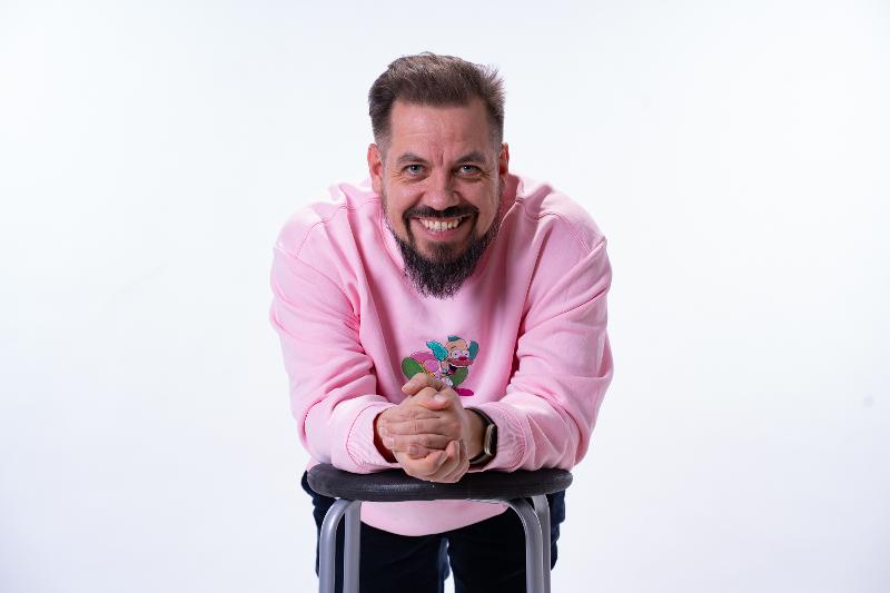 Ein mann poserar i eit kvitt studio. Han er lenar seg frmover og smilar.