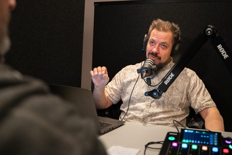 Ein mann med headset snakkar og gestikulerar foran ein mikrofon.