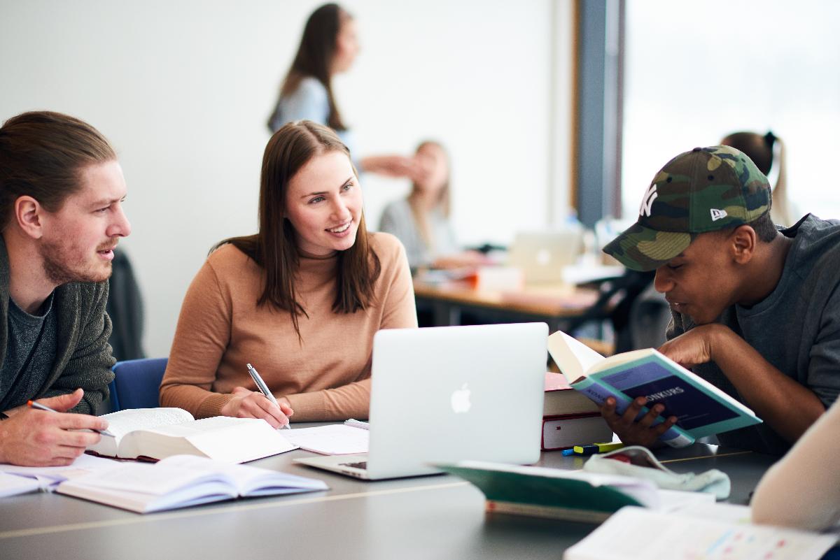 Tre unge mennesker rundt et bord, med bøker og PC