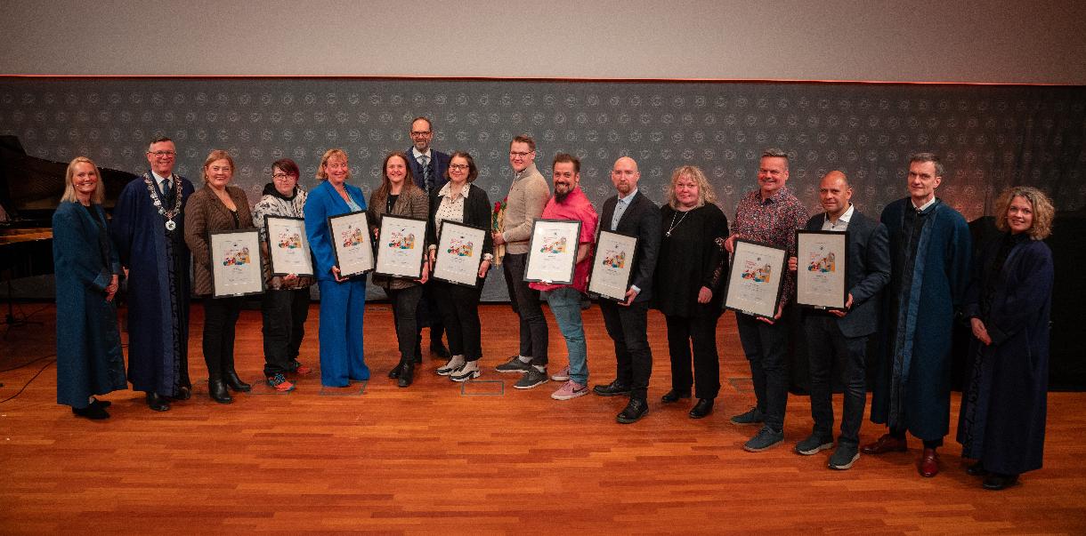 Prisvinnerne-06328.jpg En samling med prisvinnere med diplomer på høytidelig podium.