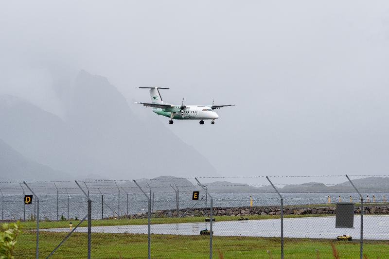 Et hvitt og gr&oslash;nt fly inn for landing i ruskev&aelig;r. Oppr&oslash;rt sj&oslash; og fjell i bakgrunnen.