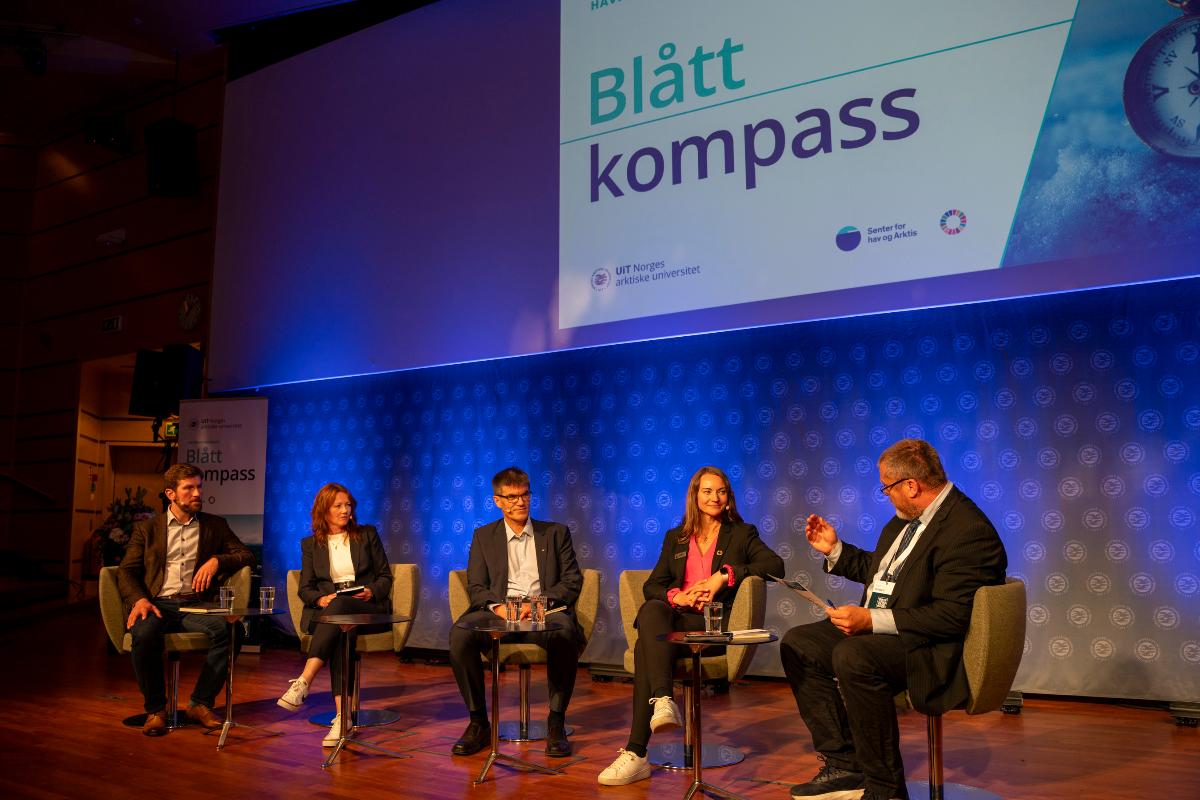 Fem mennesker sitter i stoler p&aring; en scene og diskuterer. P&aring; veggen bak dem en slide det st&aring;r Bl&aring;tt kompass p&aring;.