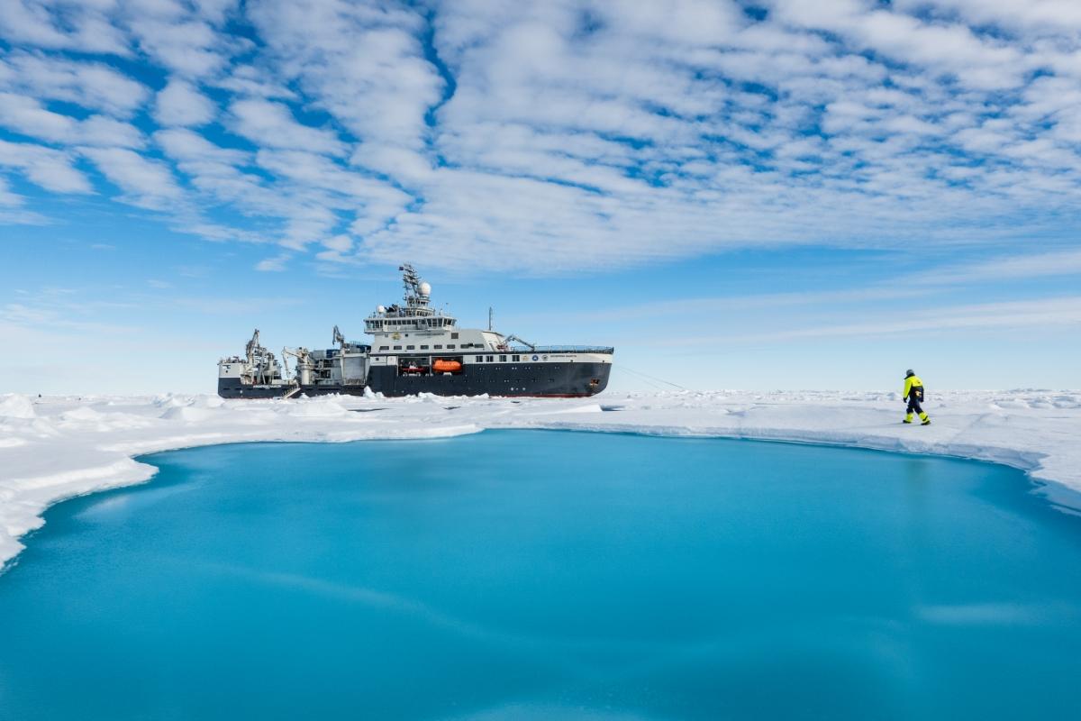 Et forskningsskip i isdekket farvann med en person i vinterutstyr n&aelig;r en &aring;pen r&aring;k. Bakgrunnen viser bl&aring; himmel og skyer. 