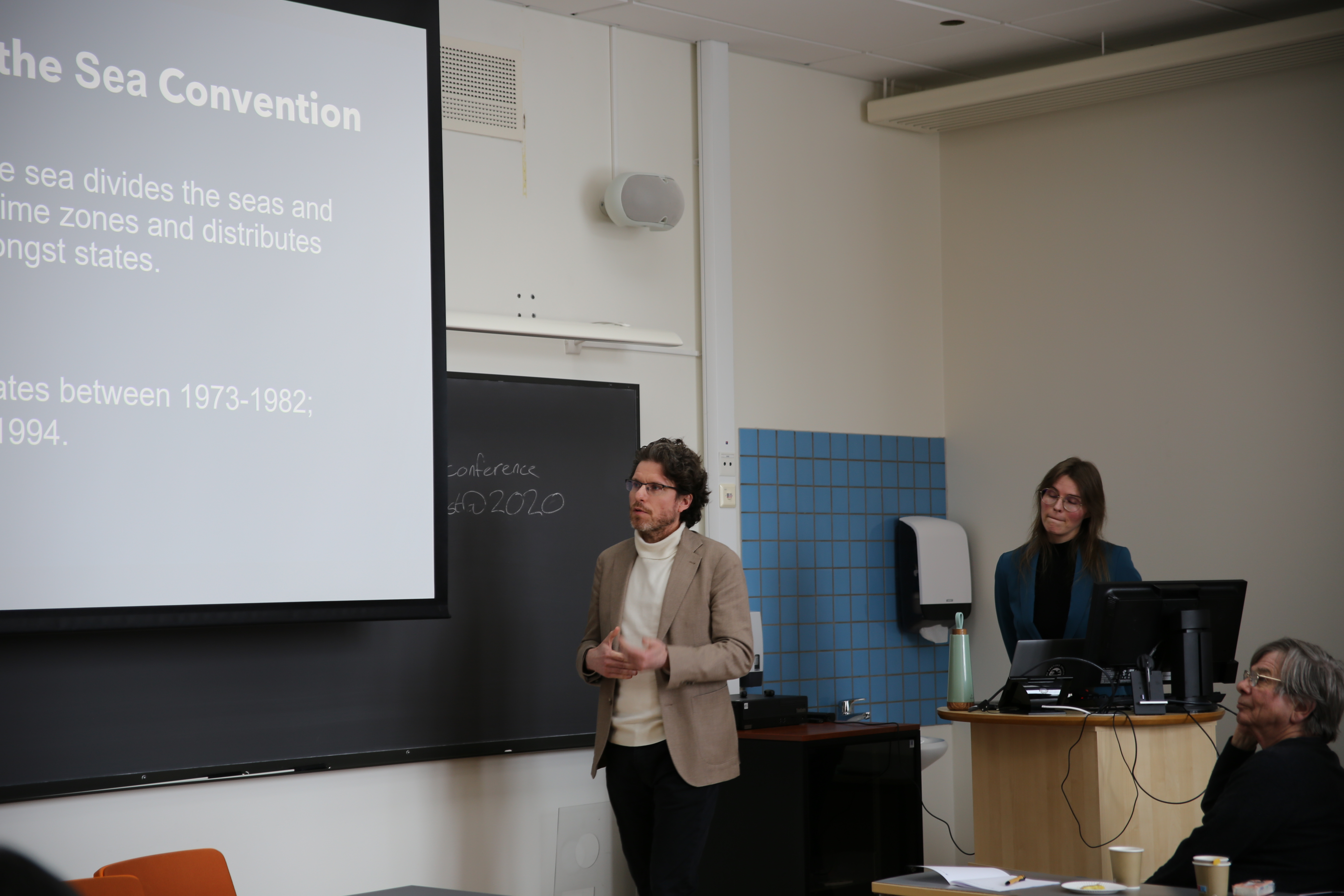 Youri speaking in front of an audience while Jessica is liostening in the backgorund