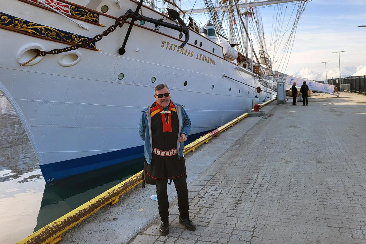 Mann ikledd en kofte st&aring;r foran en stor seilskute. 