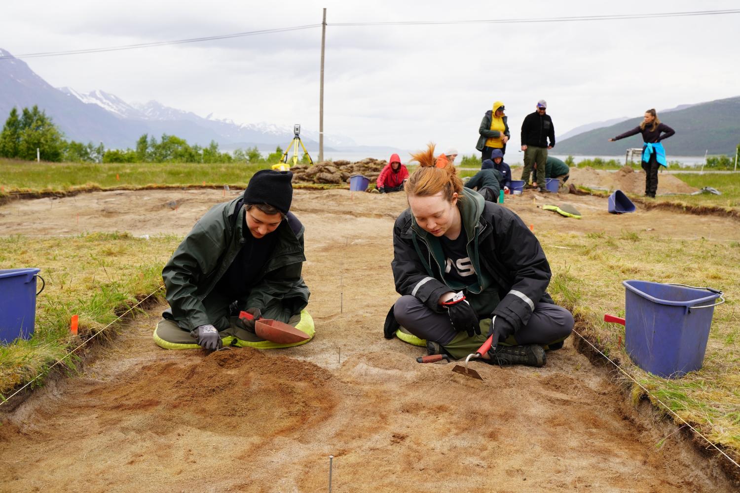 En mann og en kvinne som studerer arkeologi, sitter på bakken med utgravingsverktøy, mens fjell og andre personer er i bakgrunnen.
