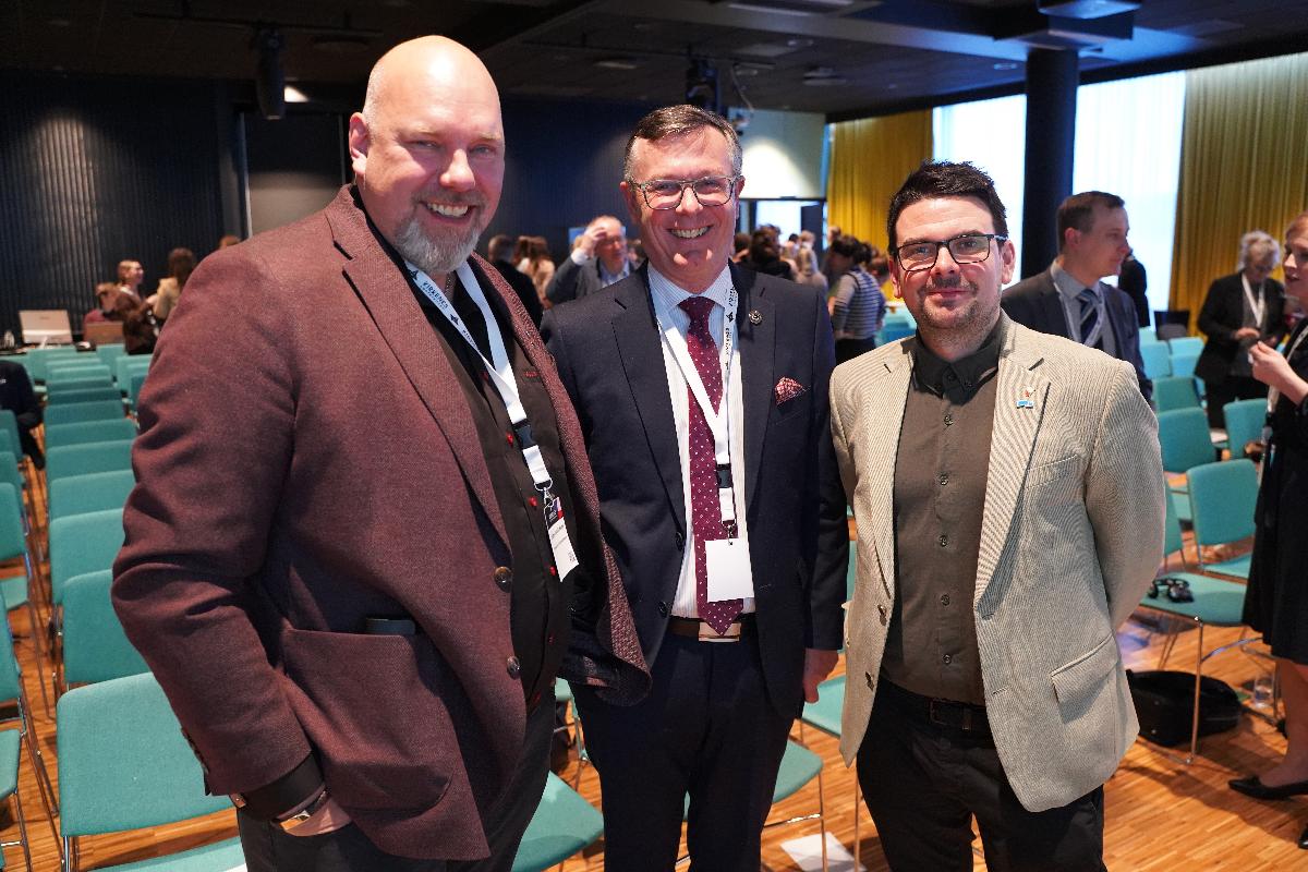 Tre menn st&aring;r ved siden av hverandre i en konferansesal