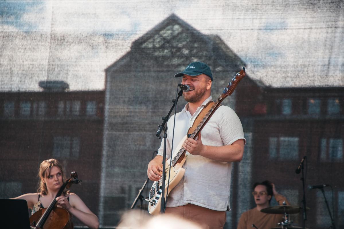 Mannlig vokalist med caps og gitar p&aring; campus-scene 