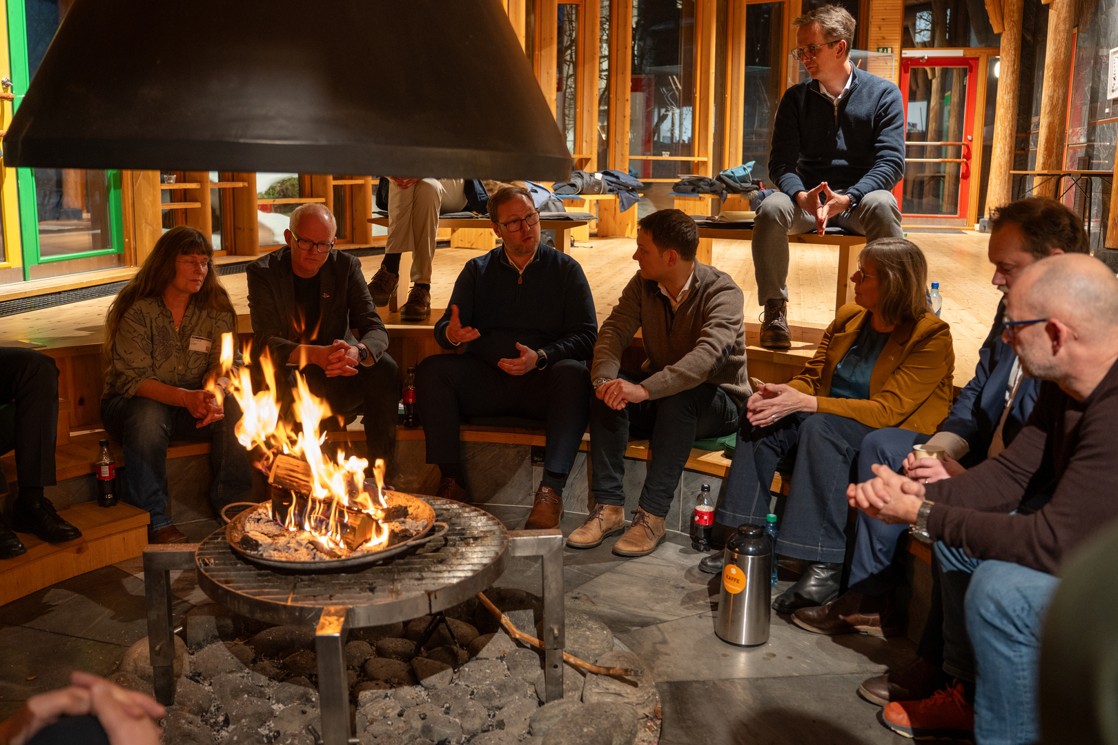 Arktisk_delegasjon_09027.jpg Personer samlet rundt et bål, innendørs.