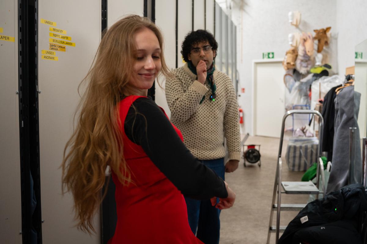 Jente med langt lyst h&aring;r svinger seg i en r&oslash;d kjole i en gang p&aring; kostymelager. Bak st&aring;r med ung mann med m&oslash;rkt h&aring;r og briller og ser p&aring; henne.