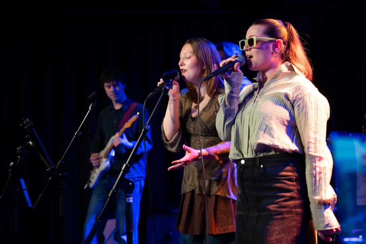 To kvinner synger, mens en mann spiller gitar i bakgrunnen. 