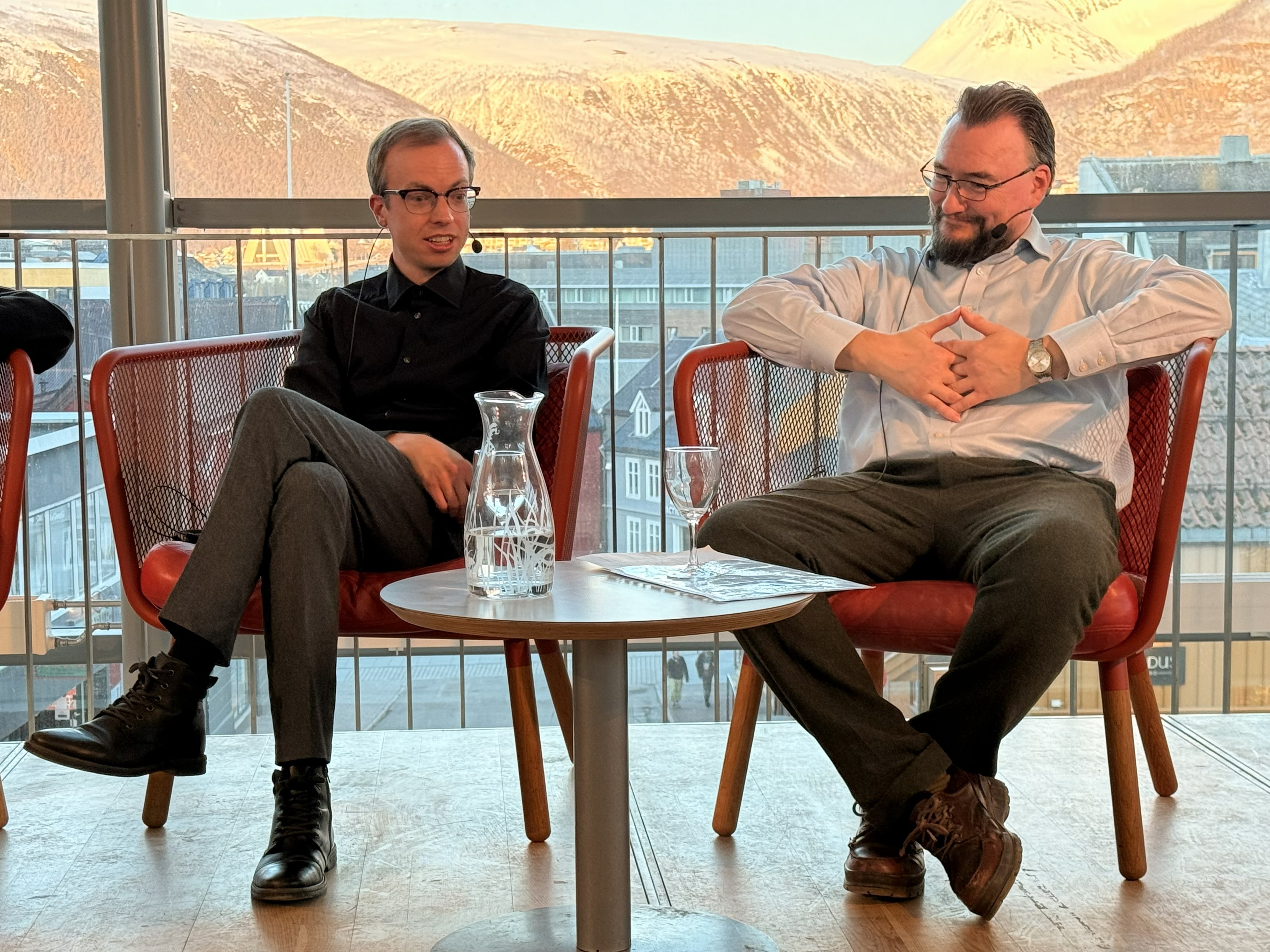 man sitting in a chair, wearing glasses, next to him a man, wearing glasses, also in a chair, large windows are in the background