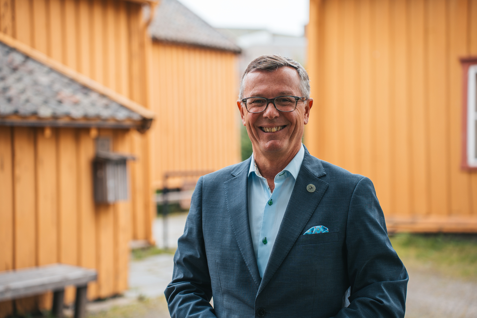 Dag Rune Olsen.jpg Rektor i dress i forgrunnen av eldre bebyggelse i oransje farge.